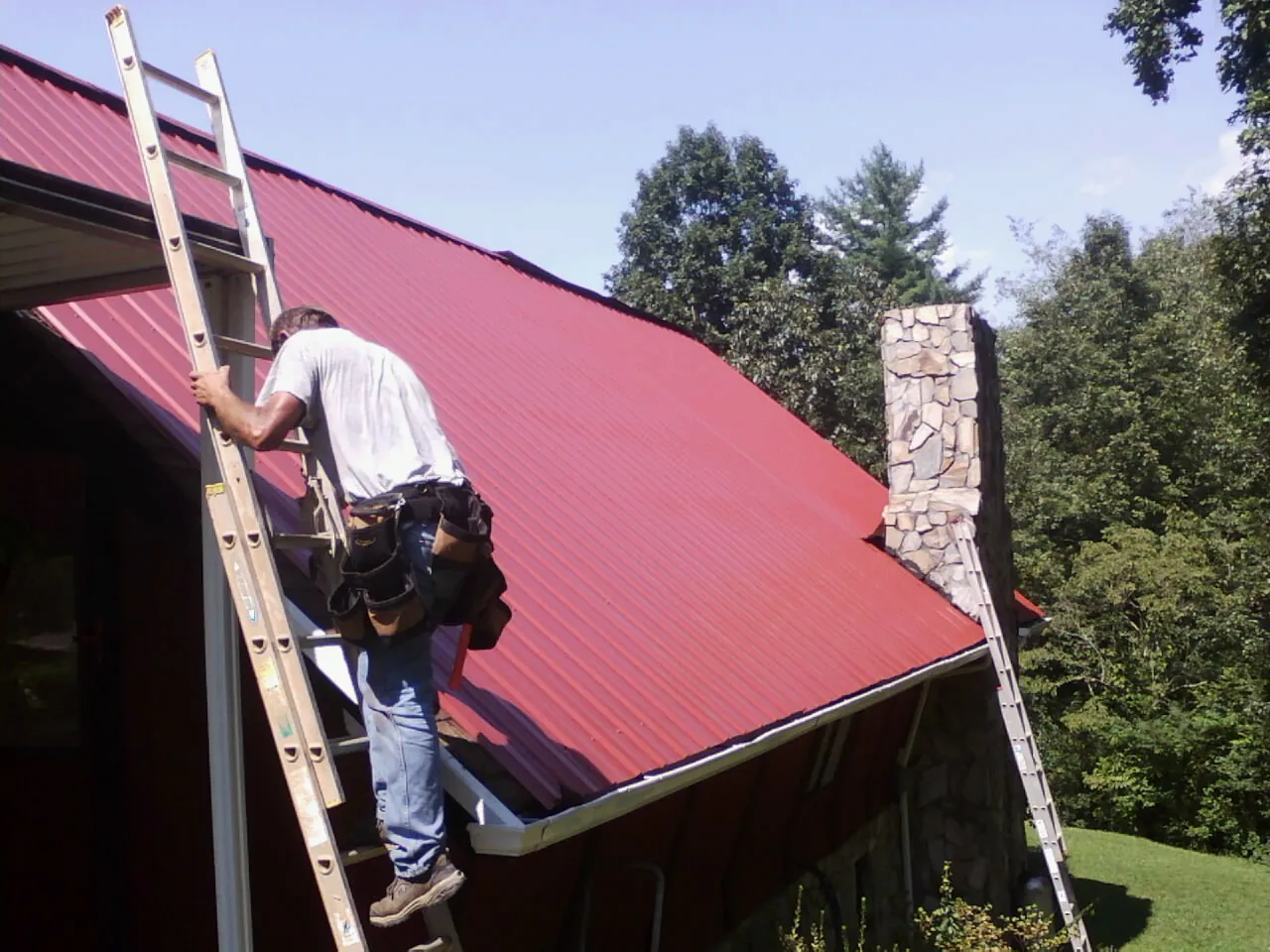 Roofer on a ladder beside a red metal roof and stone chimney — Soffit & Fascia Repair by Roofing Brothers in Sanborn